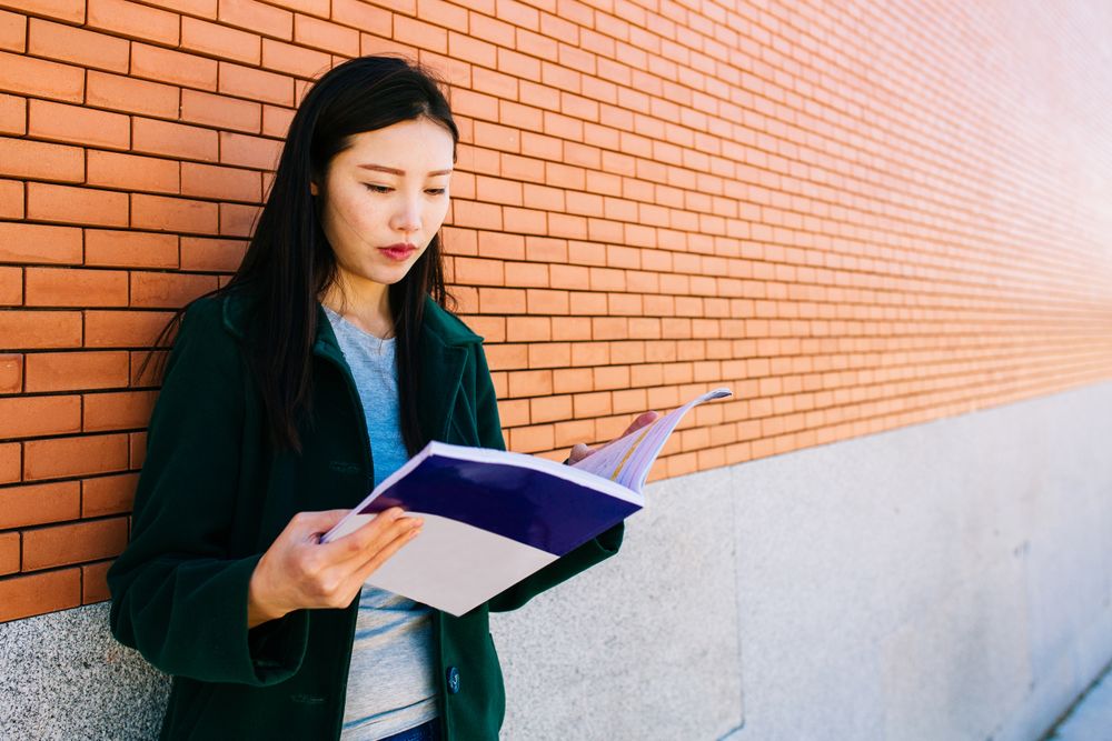 Asian student reading textbook near brick wall