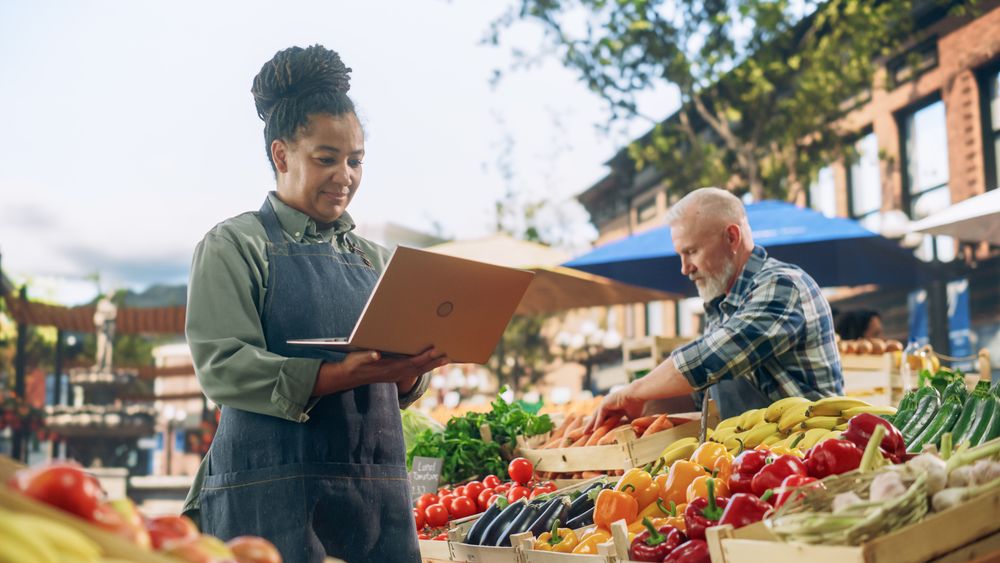 Black Farmers Market Manager Using Laptop Computer on a City Square with Vendors Selling Fresh Natural Agricultural Products.