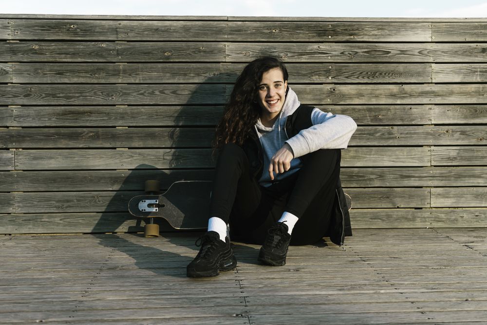 Teenage girl sitting on a longboard, leaning against a wooded wall