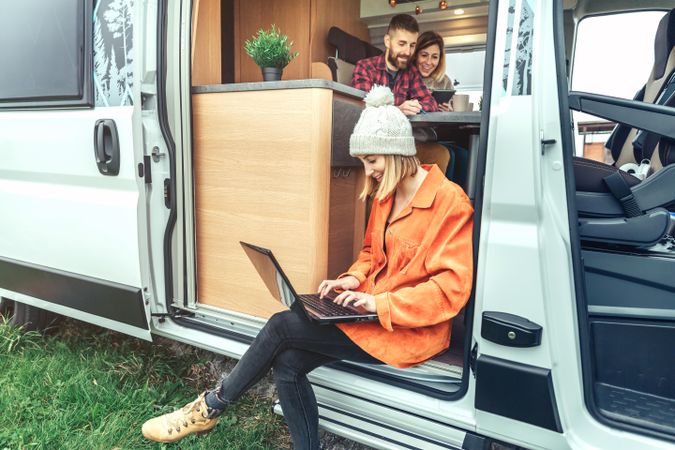 Female in jacket and woolen hat sitting on step of van with laptop