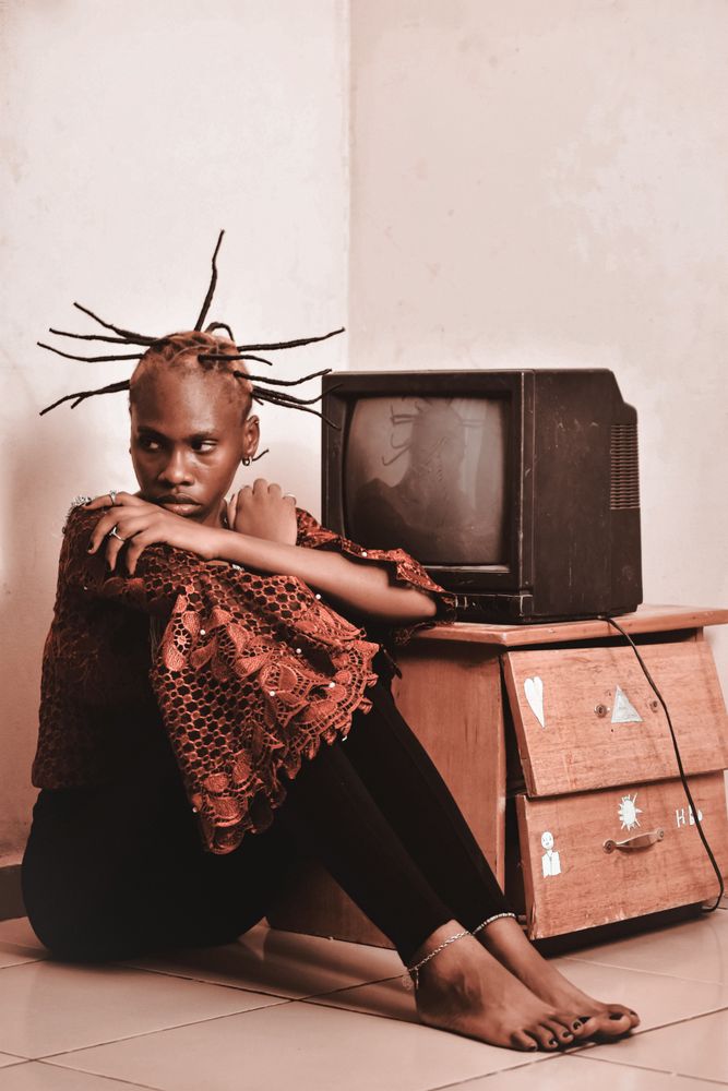 Woman with unique hairstyle wearing brown scarf sitting on floor beside crt tv