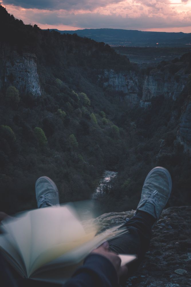 View to small river in canyon and legs of person with book sitting on the edge