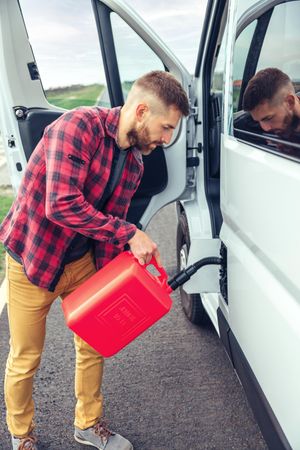 Male in red checkered shirt filling up petroleum in van with gas can, vertical