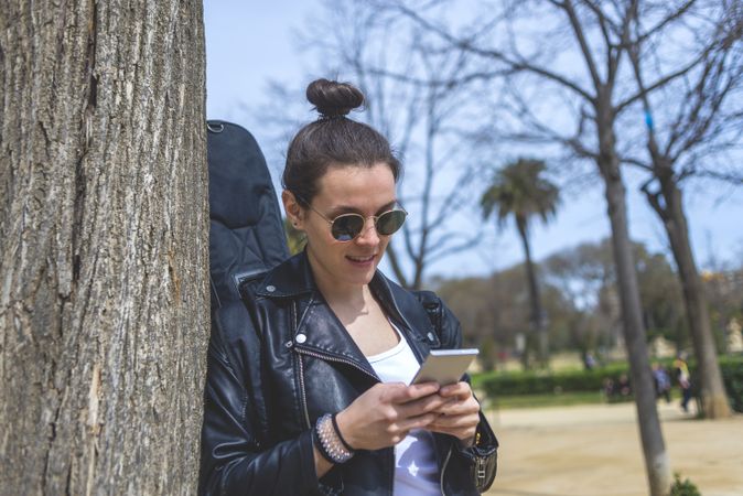 Side view of a young laughing hipster woman standing and leaning on a tree at park in sunny day while using a mobile phone