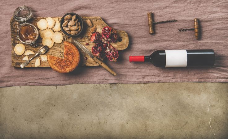 Cheese plate with bottle of red wine, cork screws, on light maroon linen, over concrete, copy space