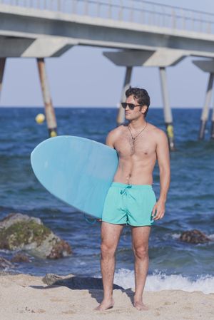 Male surfer standing in the rocks on the coast near pier, vertical