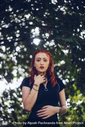 Young red-haired woman in makeup with tattoos looking at camera under tree with blurred background bGRd24