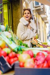 Gleeful young woman in cozy coat standing by fruits in shop