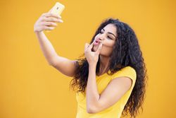 Female in yellow t-shirt taking selfie making funny face in front of mustard background