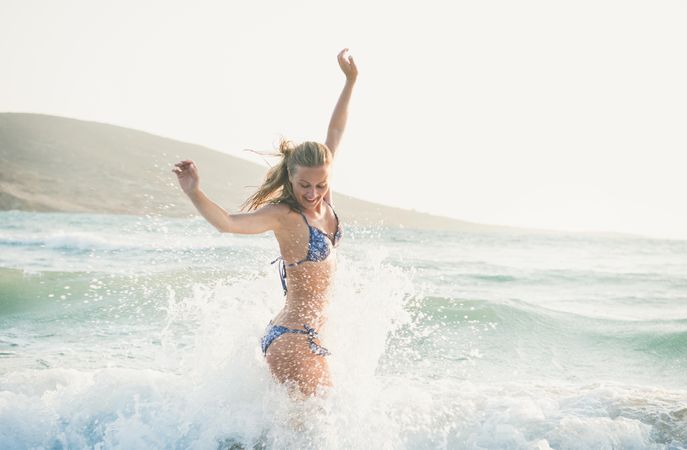 Side shot of woman in bikini jumping with outstretched arms and smiling in the water at the beach