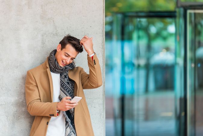 Happy man leaning on wall outside texting and wearing a camel coat