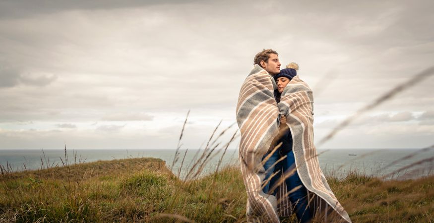 Portrait of young couple embracing under blanket in a cold day with sea and dark cloudy sky in the background