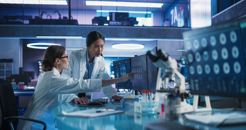 Medical Research Center Laboratory: White And Asian Female Scientists Talking And Using Desktop Computer With MRI Scans Of Human Brain.
