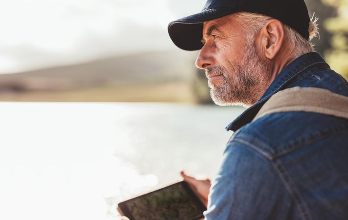 Close up portrait of mature man wearing cap sitting at a lake and looking at a view