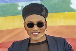 Young Latino gay man with make-up on wearing a fashionable hat and sunglasses, isolated on a Rainbow flag background. LGBT.