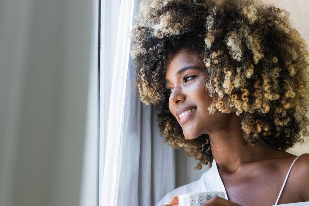 Dreamy Black woman with cup of drink near window