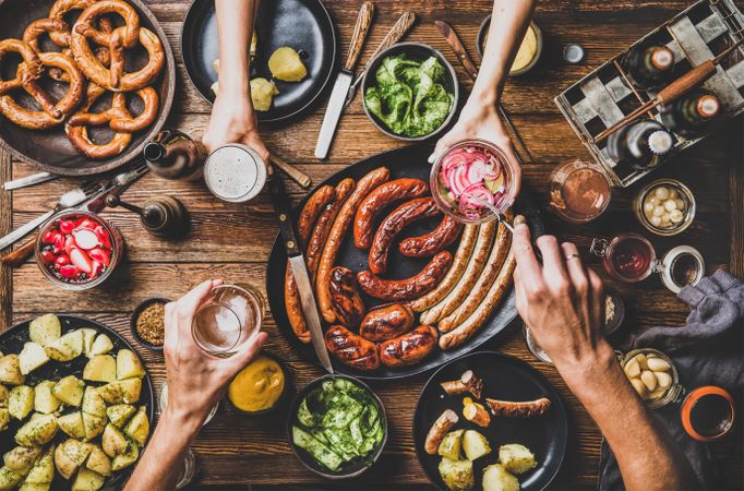 German sausages and pretzels displayed on wooden table with hands holding beer and reaching for food