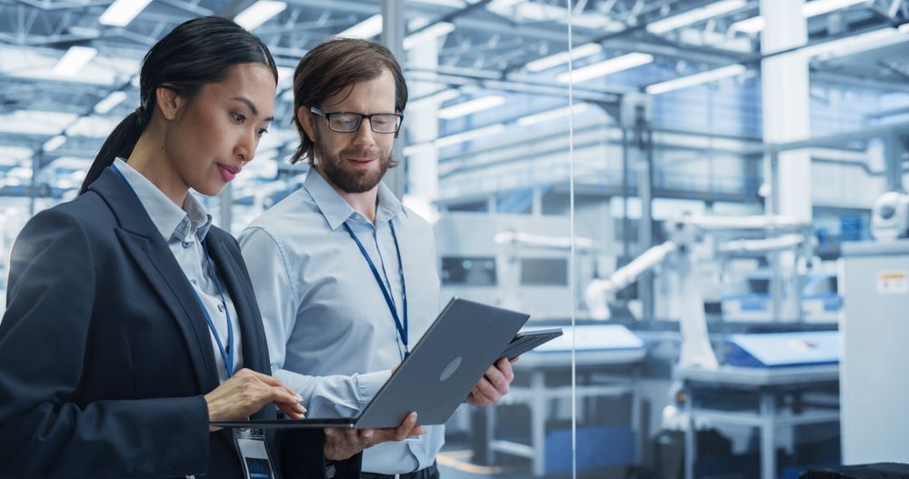 Portrait of Experienced Male and Female Engineers Standing in Agency Room, Using Computer Devices and Discussing Production at a Modern Automated Electronics Manufacture with Robot Arms