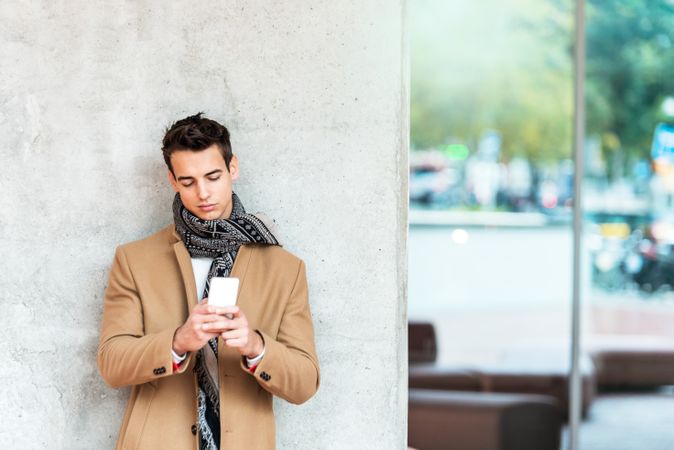 Coy man leaning on wall outside texting on phone and wearing a camel coat with copy space