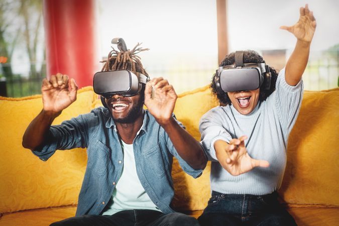 Couple of happy friends enjoying a VR game wearing goggles sitting on a yellow sofa