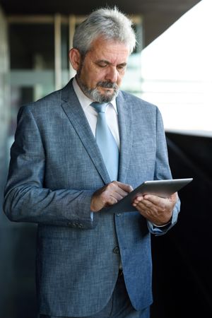 Older man in formal suit checking tablet next to grey brick wall