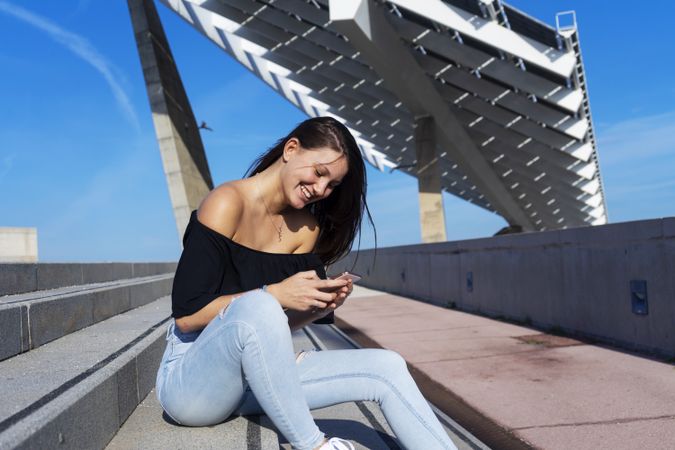 Young smiling woman with long hair sitting on promenade against harbor and checking phone