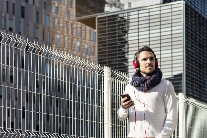Young man walking in scarf past metallic fence and looking up from smartphone with copy space