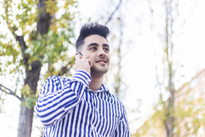 Portrait of young man standing in striped shirt speaking on phone outside