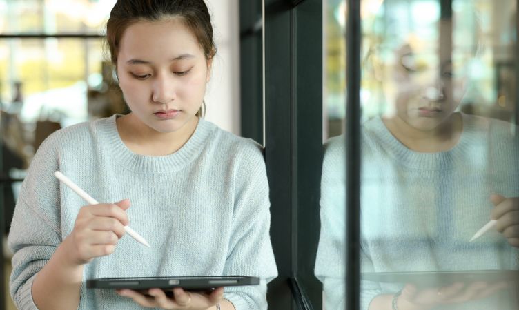 A woman holding a tablet with a pen, leaning against a glass wall.