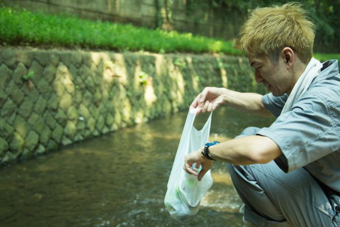 Man in work suit holding a plastic bag and crouching beside stream river