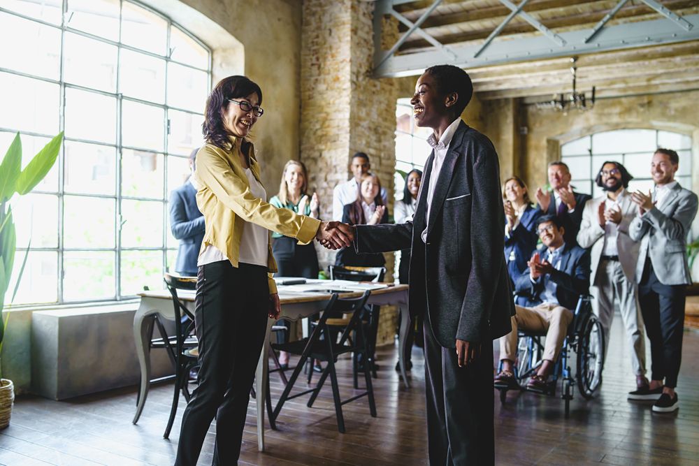 Two business women, shake hands surrounded by a multi-ethnic group of colleagues
