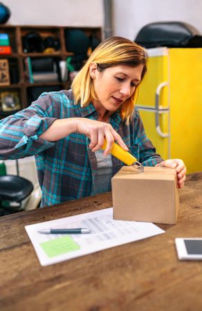 Blonde woman with cutter unpacking order received by courier with merchandise to sell on line