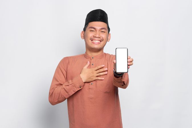 Muslim man in kufi hat smiling and presenting blank screen on mobile phone with hand on chest