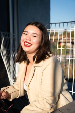 Young woman with straight brown hair sitting outside laughing with eyes closed