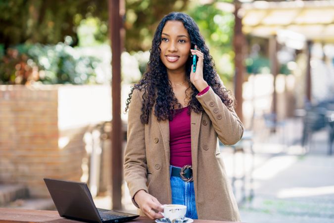 A young woman sits outdoors, using her smartphone and laptop in a workspace setting