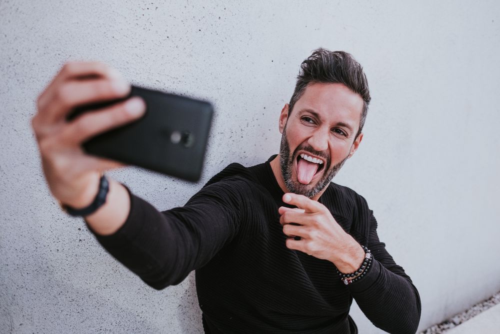 Adult stylish smiling man taking selfie on smartphone near grey wall