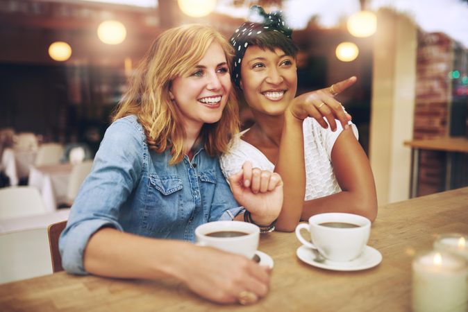 Two female friends sitting at a cafe table chatting and pointing at something outside