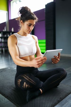 Fit woman cross legged on yoga mat reading a digital tablet, vertical