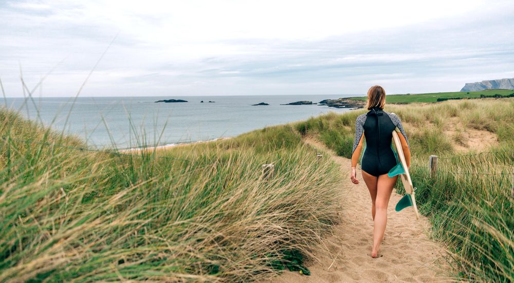 Woman walking on sandy path towards the beach holding a surfboard