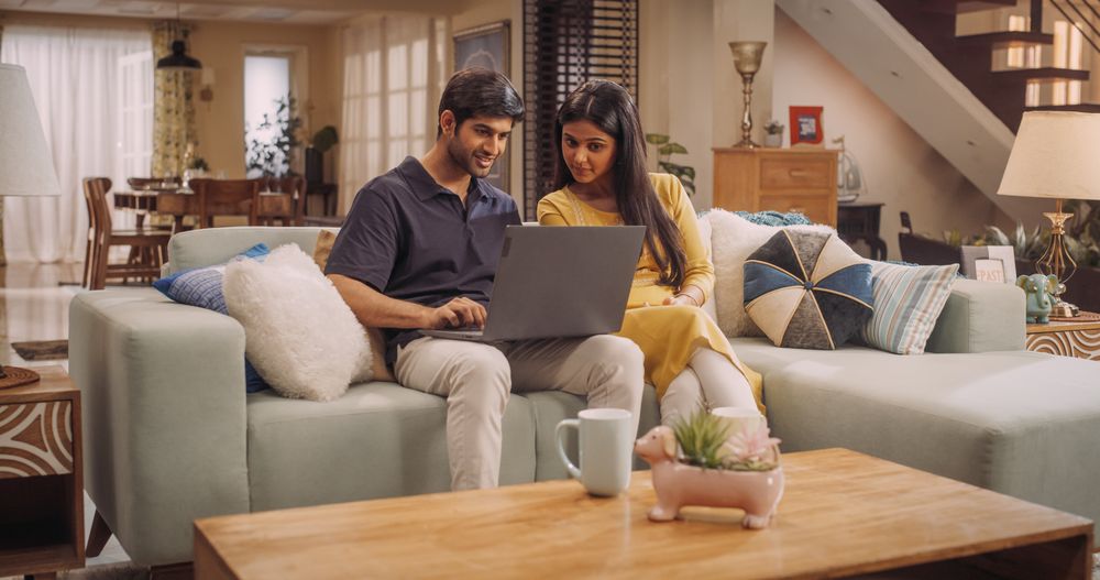 Lovely Young Indian Couple Spending time Together, Using Internet On Their Laptop, Sitting On The Sofa In Their Home.