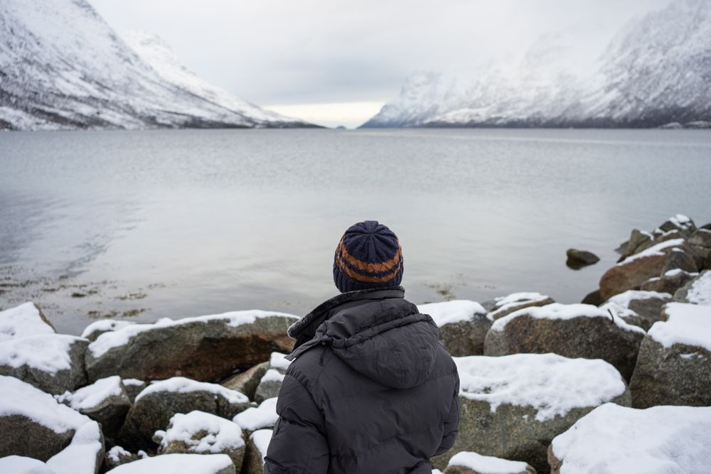 Solitary tourist on rocky coast against calm lake water and snowy mountains in cold overcast weather
