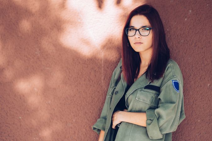 Stylish confident bright woman in glasses leaning on wall and looking at camera