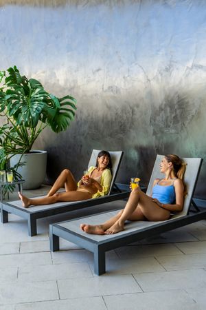 Two women friends are resting on lounge chairs, one of them holding a drink. The scene is relaxed and casual