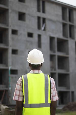 Back view of man wearing green vest and bump cap standing beside a building under construction