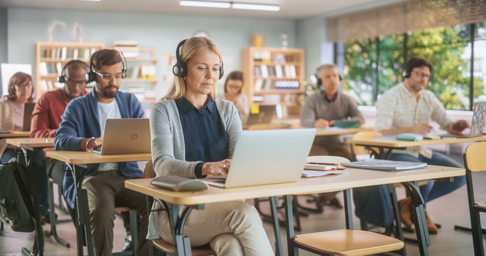 Diverse Mature Students Learning in Classroom, Sitting Behind Desks with Headphones, Using Laptops and Writing in Notebooks.