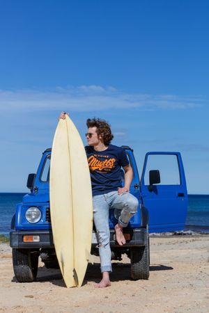 Guy with surfboard barefoot leaning on car on beach