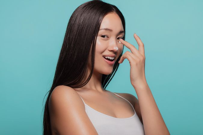 Woman applying moisturizer cream on her face against blue background