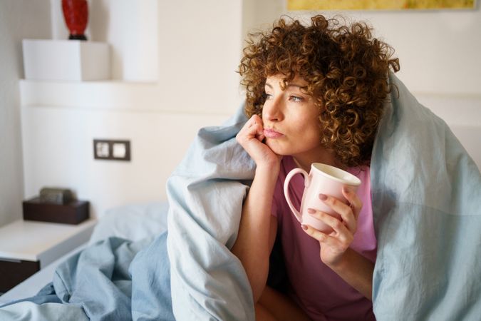 Happy woman relaxing under blue sheet in bed with cup of coffee