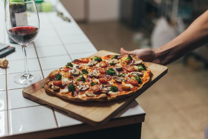 Arm laying pizza on wooden board of kitchen counter with red wine