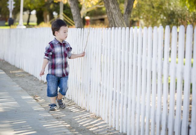 Biracial Boy Walking with Stick Along White Fence
